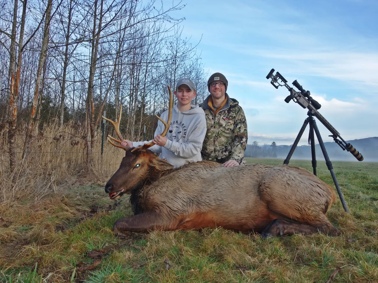 Two people with elk and rifle outdoors.