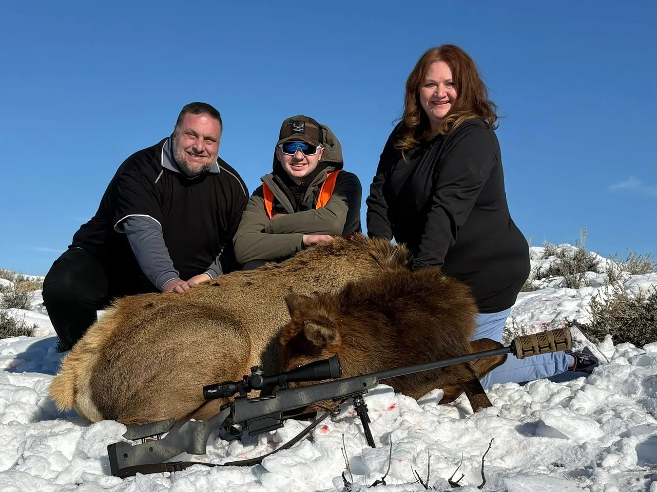 Three people posing with hunted elk outdoors.