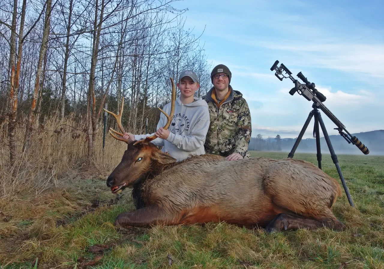 Two people with elk and rifle outdoors.