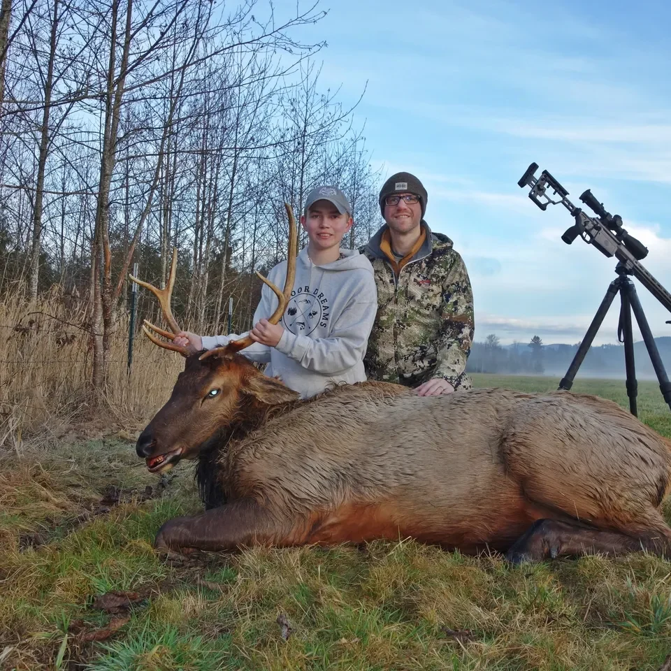Two people with elk and rifle outdoors.