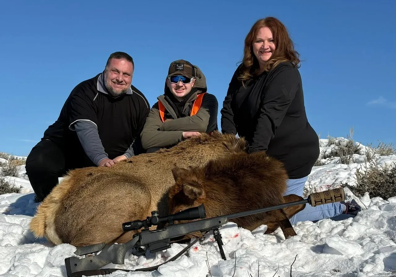 Three people posing with hunted elk outdoors.