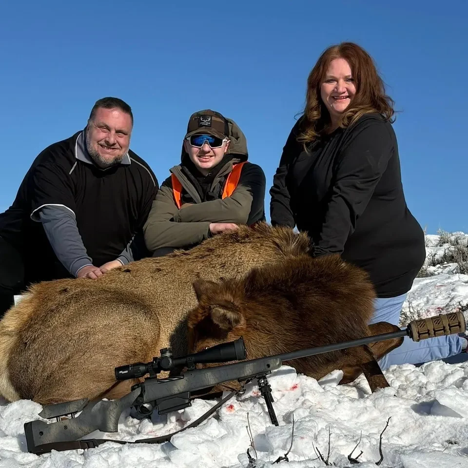 Three people posing with hunted elk outdoors.