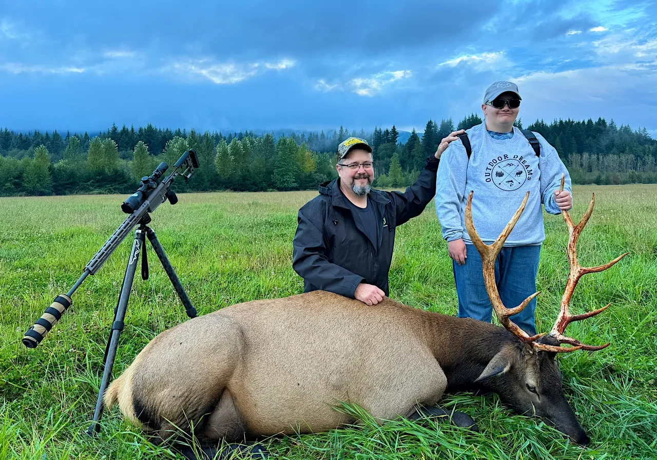 Two people pose with an elk outdoors.