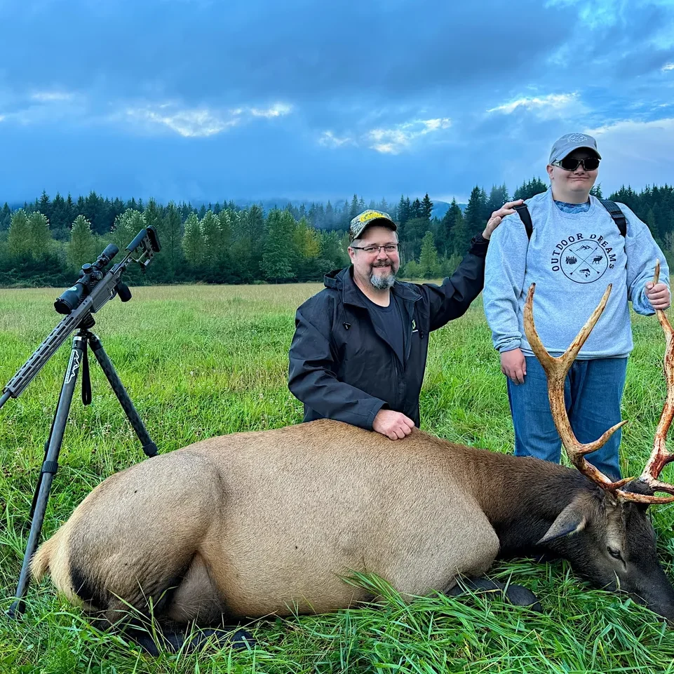 Two people pose with an elk outdoors.